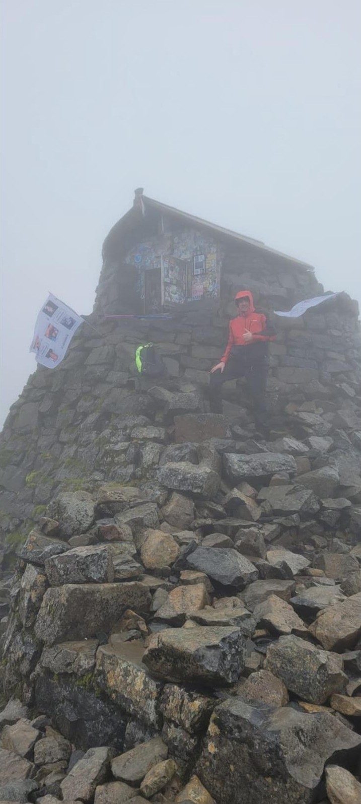 A walker at a stone summit shelter in the mist